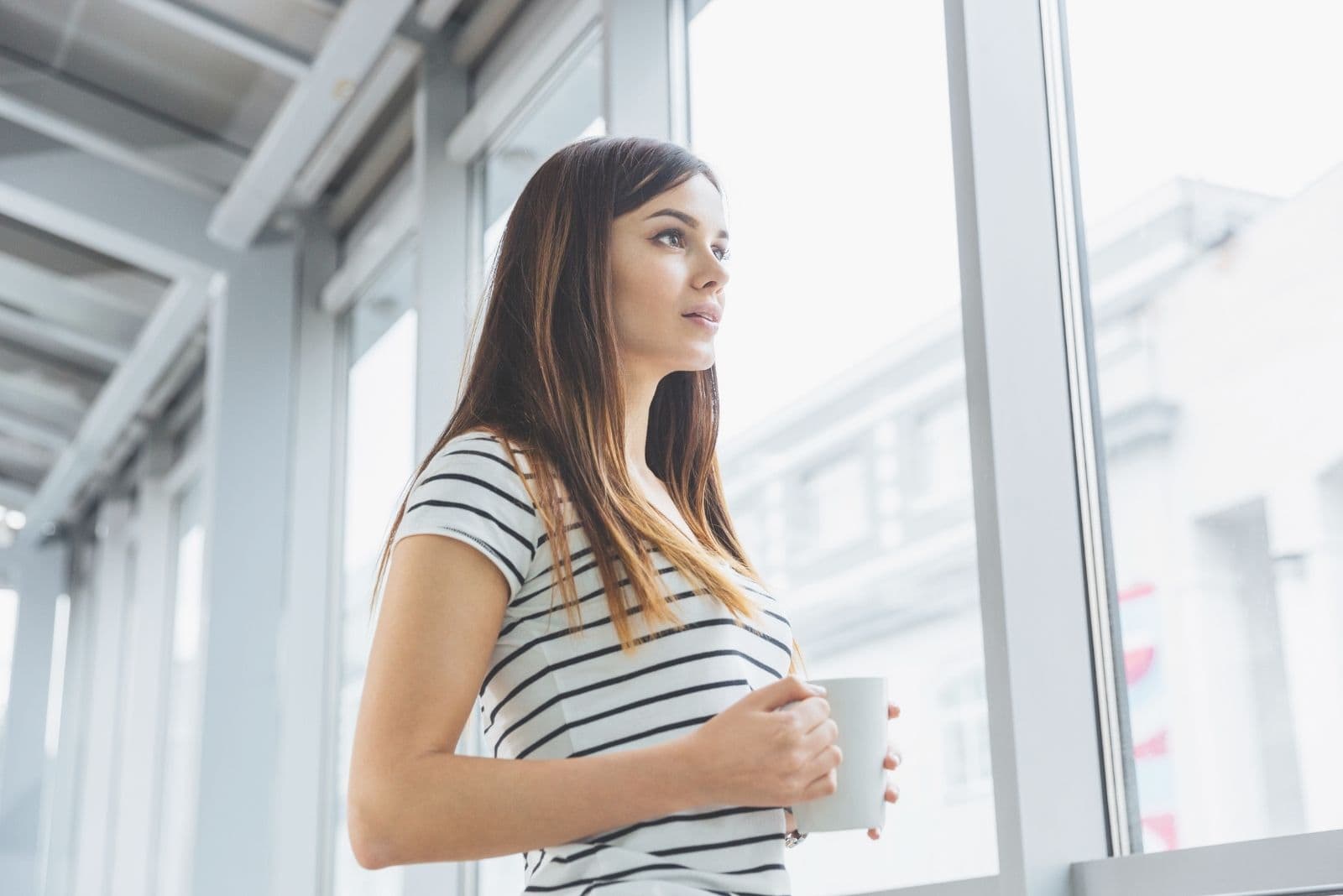 attractive pensive female holding a cup looking outside the window image in low angle